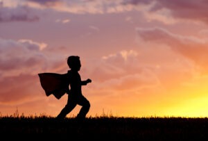 Being your own hero. Silhouette Of A young boy wearing a cape plays a super hero at sunset.
