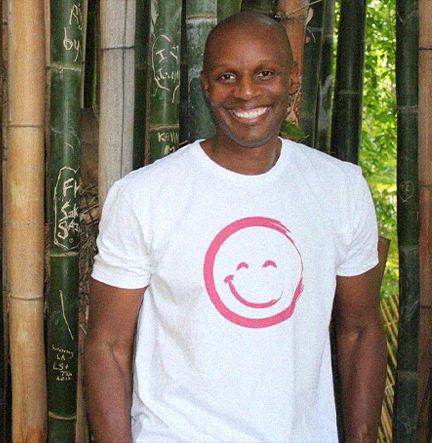 Richard Shola Smiling And Wearing a Smiley Face T-Shirt While Standing In Front of Bamboo