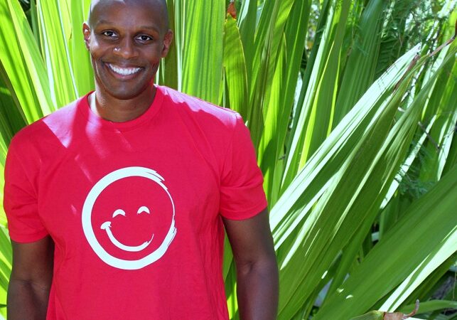 Richard Shola Smiling And Wearing a Red Smiley Face T-Shirt While Standing In Front of Large Plant